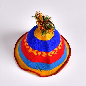 May include: A colorful, handmade hat with a wide brim. The hat features horizontal stripes in red, yellow, and blue, with a decorative pattern in yellow. A tassel of yellow, orange, and green yarn adorns the top. The hat is likely made of yarn or a similar material.