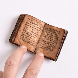 May include: A miniature open book with aged, tan-colored parchment pages featuring dark, hand-written Ge'ez script. The small volume has visible binding along the spine and shows signs of wear on the paper edges, displaying dense text.