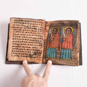 Puede incluir: Un manuscrito antiguo abierto con páginas de pergamino beige envejecido. La página izquierda presenta una densa escritura negra en ge'ez. La página derecha muestra una colorida ilustración de dos figuras aladas con túnicas rojas y faldas azul verdoso.