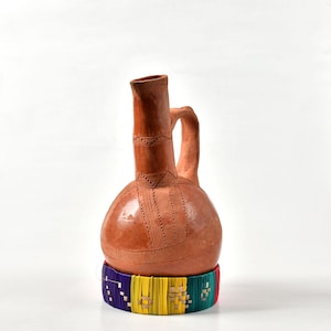 May include: A terracotta-colored ceramic jug with a handle and a decorative band at the base. The jug has etched patterns and a colorful woven band in purple, yellow, green, and blue. The jug is likely used for serving beverages.
