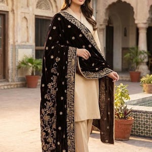 Puede incluir: Una mujer con un conjunto beige y un chal de terciopelo marrón oscuro con bordados dorados y diseños florales. El conjunto se presenta en un patio exterior con plantas en macetas.