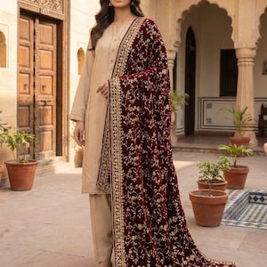 Puede incluir: Una mujer con un salwar kameez beige y un chal de terciopelo burdeos. El chal está adornado con bordados florales dorados. El conjunto se completa con bailarinas doradas. El fondo presenta un entorno al aire libre con detalles arquitectónicos.