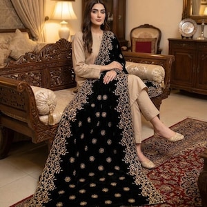 May include: A black velvet shawl with intricate gold embroidery. The shawl is draped over a person wearing a beige outfit. The design features floral and geometric patterns. The person is seated on a carved wooden bench.