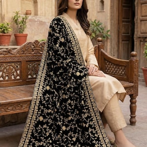 May include: A woman wearing a beige outfit with a black shawl. The shawl is embroidered with gold floral patterns. She is sitting on a wooden bench in an outdoor setting.
