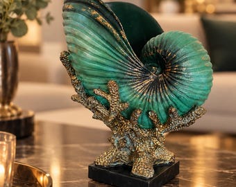 Puede incluir: Una escultura decorativa que presenta una concha de nautilo de color verde azulado con reflejos dorados, apoyada sobre una base texturizada similar al coral. La pieza está montada sobre un pedestal cuadrado de mármol negro.