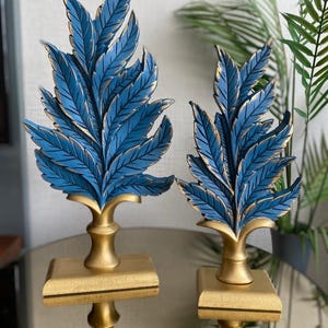 Könnte beinhalten: Zwei dekorative Blattskulpturen. Jede Skulptur zeigt eine Ansammlung blauer Blätter mit goldenen Akzenten, die auf einem goldenen Sockel montiert sind. Die Skulpturen werden auf einer reflektierenden Oberfläche präsentiert.