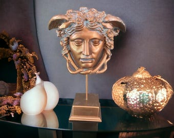 Puede incluir: Una escultura decorativa dorada de una cabeza de Medusa con serpientes alrededor de su cabeza, sobre un pedestal dorado. La escultura está sobre una superficie de vidrio verde oscuro.
