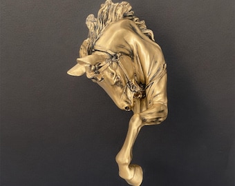 May include: A gold-colored horse head sculpture with a raised leg. The horse's head is tilted down and the sculpture is mounted on a wall.