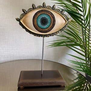 Op de afbeelding: Een decoratief oogbeeldhouwwerk met een lichtbeige iris, een turkooizen pupil en een zwart centrum. Het oog is omlijst door een goudkleurige, sierlijke rand en gemonteerd op een metalen staaf die aan een bruine rechthoekige basis is bevestigd.