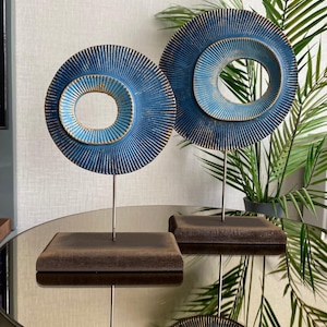 May include: Two blue and gold decorative sculptures. Each sculpture features a circular design with concentric rings and a textured surface. They are mounted on a dark brown rectangular base and stand on a reflective surface.