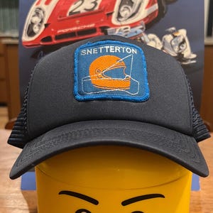 May include: A black trucker hat with a blue and orange patch that says "Snetterton" and depicts a racing helmet. The hat is sitting on top of a yellow Lego head with a smiling face.