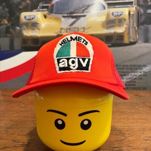 May include: A red trucker hat with a white, green, and red patch that says "HELMETS AGV." The hat is on top of a yellow Lego head. The background features a vintage racing poster.