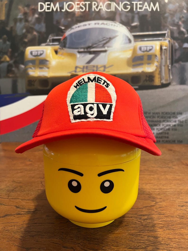 May include: A red trucker hat with a white, green, and red patch that says "HELMETS AGV." The hat is on top of a yellow Lego head. The background features a vintage racing poster.