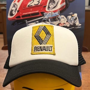 Op de afbeelding: Een witte en zwarte trucker-pet met een gele ruitvormige patch waarop "RENAULT" staat. De pet zit op een geel Lego-hoofd met een zwarte glimlach en wenkbrauwen.