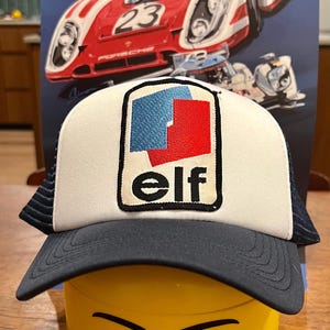 May include: A white and blue trucker hat with a red and blue "elf" logo patch. The hat is sitting on top of a yellow Lego head with a smiling face.