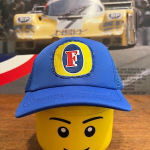 May include: A blue and yellow trucker hat with a large white letter "F" on the front. The hat is sitting on top of a yellow Lego head with a smiling face.