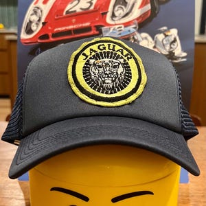 May include: A black and yellow trucker hat with a jaguar logo patch. The hat is sitting on top of a yellow Lego head with a smiling face.