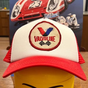 Puede incluir: Una gorra de camionero blanca y roja con un parche de logotipo de Valvoline. El logotipo es una V roja, blanca y azul con la palabra Valvoline en letras blancas y un diseño de bandera a cuadros debajo. La gorra está sentada encima de una cabeza de Lego amarilla con una cara sonriente.
