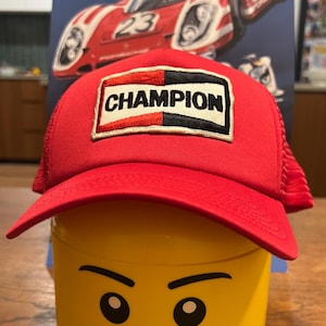 May include: A red trucker hat with a Champion patch, worn by a yellow Lego head. The Champion patch is black, white, and red. A poster of Porsche race cars is in the background.