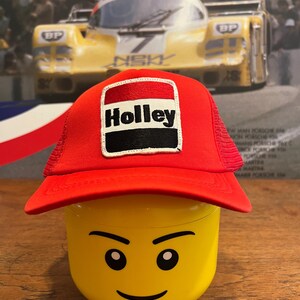 May include: A red and white trucker hat with a black and white Holley logo patch. The hat is sitting on top of a yellow Lego head.