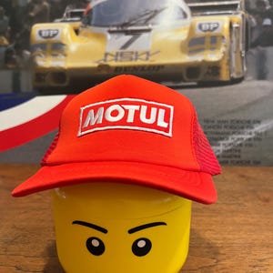 May include: A red and black trucker hat with a white "MOTUL" logo on the front. The hat is sitting on top of a yellow Lego head.