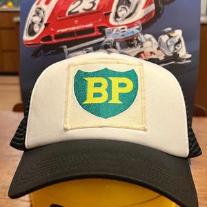 May include: A white and black trucker hat with a green and yellow BP logo patch. The hat is sitting on top of a yellow Lego head with a smiling face.