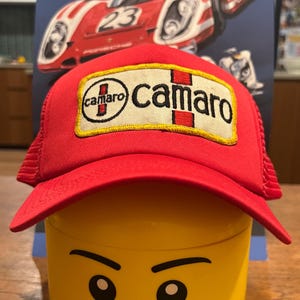 May include: Red and white trucker hat with a yellow and red embroidered patch that says "Camaro" on it. The hat is sitting on top of a yellow Lego head.