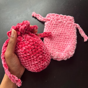May include: Two handmade, plush drawstring bags. One is held in a hand, the other rests on a dark surface. The bags are made of a soft, textured material in shades of pink and red. The drawstring cords are visible.