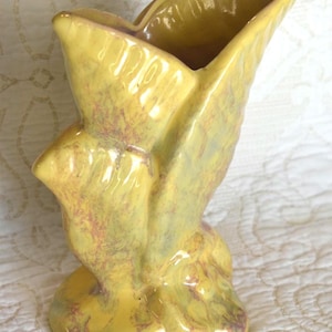 May include: A vintage yellow ceramic vase with an unusual, asymmetrical design. The vase has a flared opening and a textured surface with hints of purple and green. It stands approximately 20 cm tall.