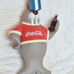 Puede incluir: Una morsa gris que lleva un suéter rojo con el logotipo de Coca-Cola y sostiene una botella de Coca-Cola. La morsa lleva una caja de regalo azul en la cabeza.