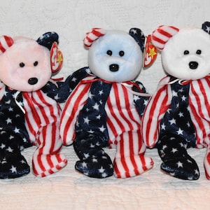 May include: Three plush teddy bears with patriotic designs. Each bear has a different colored head: pink, light blue, and white. The bears' bodies are decorated with a pattern of red and white stripes and blue stars.
