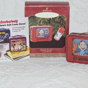 Peut inclure: Un ensemble de deux ornements Hallmark Keepsake représentant des boîtes à lunch vintage. La boîte à lunch rouge présente une image de dessin animé de Howdy Doody du spectacle Howdy Doody Show. L'autre boîte à lunch est un téléviseur rouge et bleu avec une image de dessin animé d'un garçon à l'intérieur. Le texte "Exclusively at Hallmark Gold Crown Stores!" est imprimé sur une carte blanche.