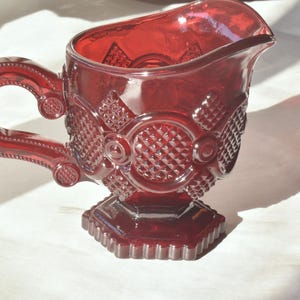 Puede incluir: Una jarra de crema de vidrio roja con un patrón de diamante y un asa. La jarra tiene una base ancha y una parte superior redondeada.