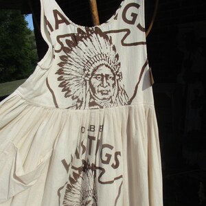 May include: A white cotton sleeveless dress with a brown print of a Native American chief wearing a headdress. The dress has a large pocket on the front and the text "Oldest" and "Hastings" printed on the front.