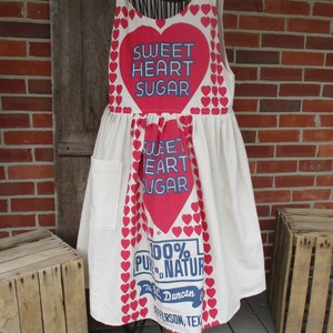 May include: A cream-colored apron with a large red heart design that says "SWEET HEART SUGAR" in blue text. The apron has a pocket and is decorated with small red hearts. The bottom of the apron has text that reads "100% PURE NATURAL".
