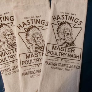 May include: Three white sacks with the text "HASTINGS" and "MASTER POULTRY MASH" printed in brown. Each sack is labeled "100 LBS. NET" and features an illustration of a Native American chief.