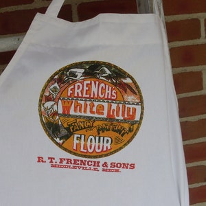 May include: White cotton apron featuring a vintage-style circular graphic with the text "FRENCH'S White Lily Fancy Patent Flour." The apron also includes the text "R.T. FRENCH & SONS, MIDDLEVILLE, MICH." in red. The apron is hanging against a brick wall.