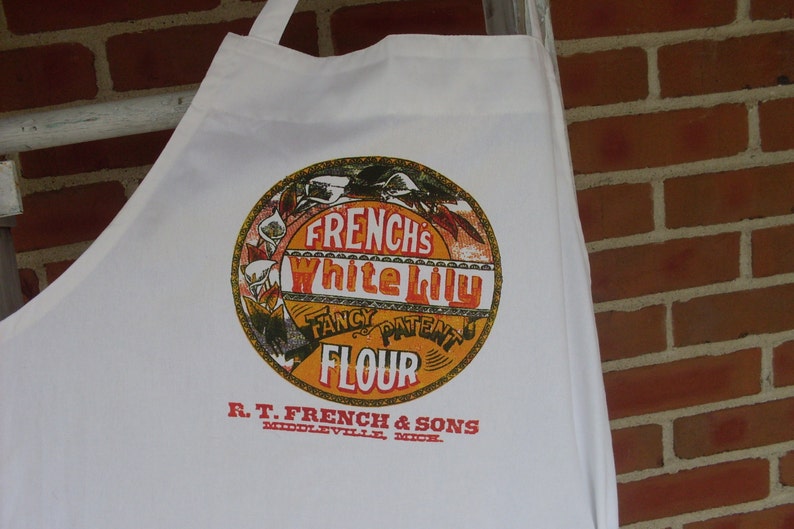 May include: White cotton apron featuring a vintage-style circular graphic with the text "FRENCH'S White Lily Fancy Patent Flour." The apron also includes the text "R.T. FRENCH & SONS, MIDDLEVILLE, MICH." in red. The apron is hanging against a brick wall.