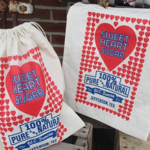 May include: Two off-white fabric bags with red and blue text and heart graphics. One is a drawstring bag, the other a tote bag. The text reads "SWEET HEART SUGAR" and "100% PURE & NATURAL" with the company name and location.