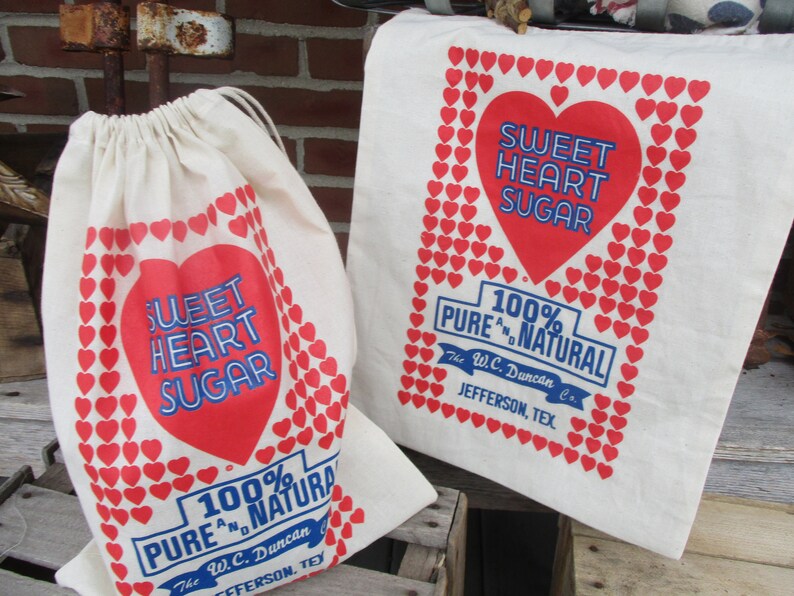 May include: Two off-white fabric bags with red and blue text and heart graphics. One is a drawstring bag, the other a tote bag. The text reads "SWEET HEART SUGAR" and "100% PURE & NATURAL" with the company name and location.