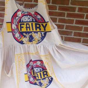 May include: A cream-colored apron made from a flour sack, featuring the text "FRENCH'S FAIRY" in a circular design with red, blue, and yellow accents. The apron has a gathered waist and a vintage aesthetic, with the words "CAKE & PASTRY FLOUR" printed on the bottom.