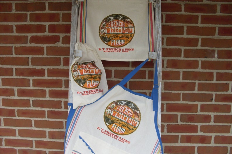 May include: A collection of vintage-style kitchen textiles, including an apron, a flour sack, and a dish towel. Each item features a circular logo with the text "French's White Lily Flour" in orange and brown, with blue and red stripes.