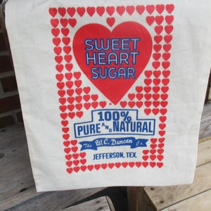 May include: A white canvas tote bag with a red heart design and the words "Sweet Heart Sugar" in blue. The bag is framed with red hearts and includes text that reads "100% Pure and Natural" and "Jefferson, Tex."