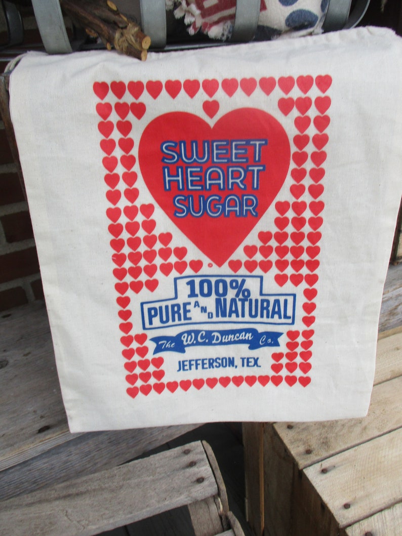 May include: A white canvas tote bag with a red heart design and the words "Sweet Heart Sugar" in blue. The bag is framed with red hearts and includes text that reads "100% Pure and Natural" and "Jefferson, Tex."