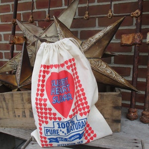 May include: A cream-colored drawstring bag with a red heart-shaped design that reads "Sweet Heart Sugar." The bag also features the text "100% Pure and Natural" and "The W.C. Duncan Co. Jefferson, Tex."