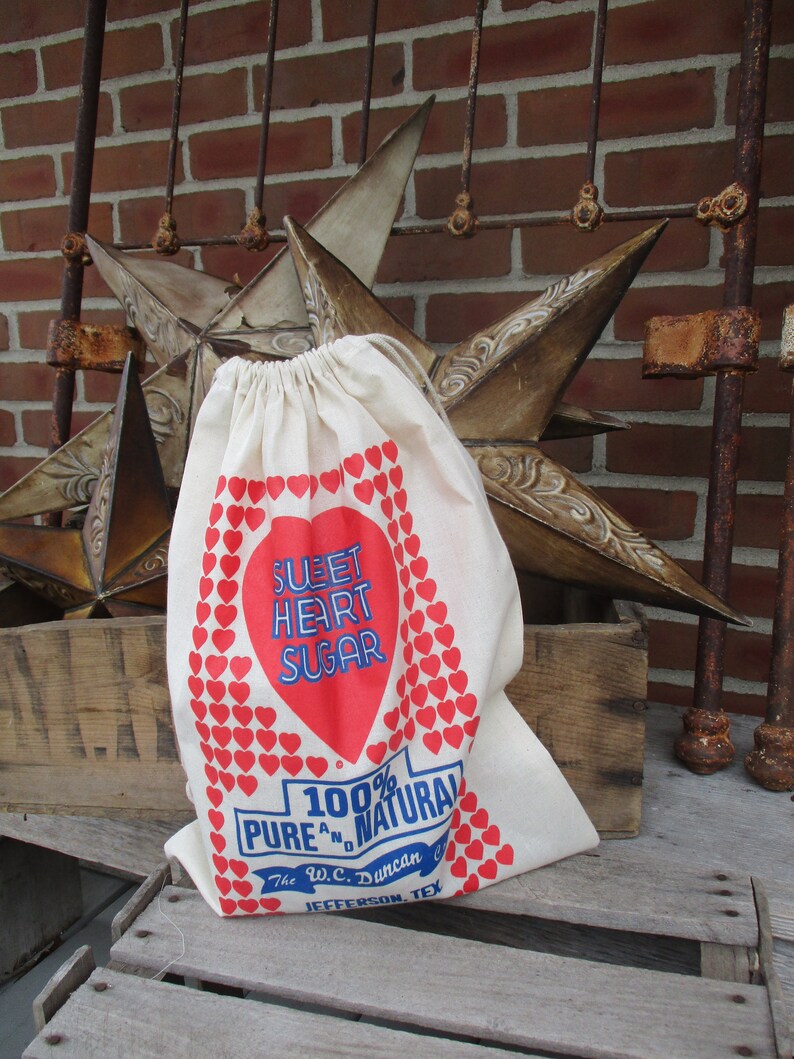 May include: A cream-colored drawstring bag with a red heart-shaped design that reads "Sweet Heart Sugar." The bag also features the text "100% Pure and Natural" and "The W.C. Duncan Co. Jefferson, Tex."