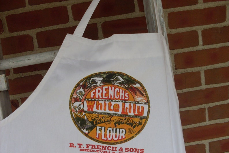 May include: White apron with a circular graphic featuring the text "FRENCH'S White Lily Fancy Patent Flour." The apron is hanging against a brick wall and a white ladder.