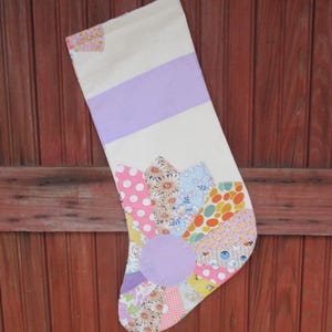 May include: A handmade Christmas stocking with a patchwork design. The stocking features a white body with a lavender stripe and a patchwork toe with floral and polka dot patterns. A small loop is attached for hanging.