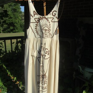 May include: A white cotton dress with brown print featuring an image of a Native American wearing a headdress. The print also includes the text "Higgins", "Peter", "Ay Mas", "with", "Concentrated", "& Bean", "Mich".