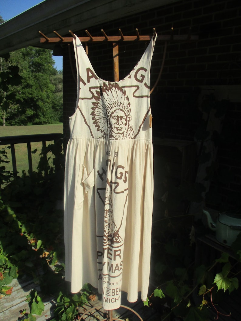 May include: A white cotton dress with brown print featuring an image of a Native American wearing a headdress. The print also includes the text "Higgins", "Peter", "Ay Mas", "with", "Concentrated", "& Bean", "Mich".